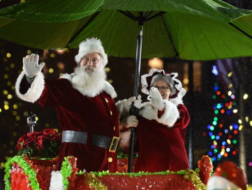 Santa and Mrs. Claus wave from a festive float, sheltered under a large green umbrella, with colorful lights in the background.