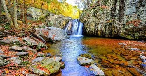 A serene waterfall cascades into a clear pool, surrounded by autumn foliage and rocky terrain.