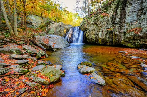 A serene waterfall cascades into a clear pool, surrounded by autumn foliage and rocky cliffs.