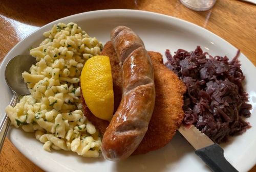 A plate with a sausage, breaded cutlet, lemon slice, spätzle pasta, and red cabbage.