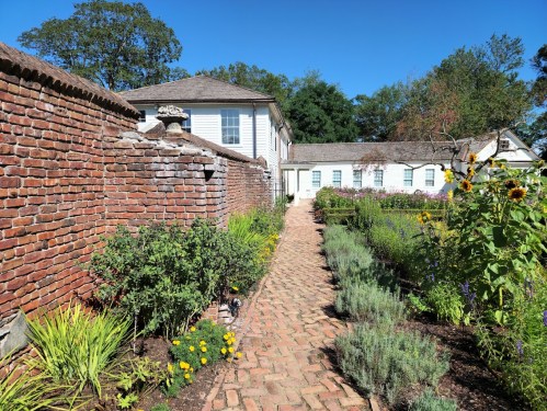 A brick pathway lined with flowers leads to a white house, surrounded by greenery and blue skies.