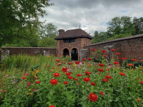 A brick building with an arched entrance surrounded by vibrant red flowers and lush greenery under a cloudy sky.
