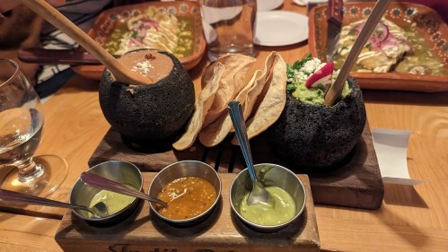 A wooden platter with two stone bowls of dips, crispy tortillas, and three small bowls of sauces.
