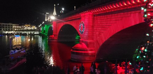 A brightly lit bridge at night, adorned with red and green lights, reflecting in the water below.