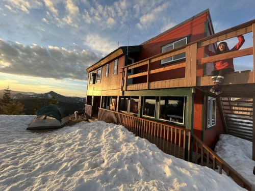 A mountain cabin with a deck, surrounded by snow, and a person standing on the balcony, enjoying the view.