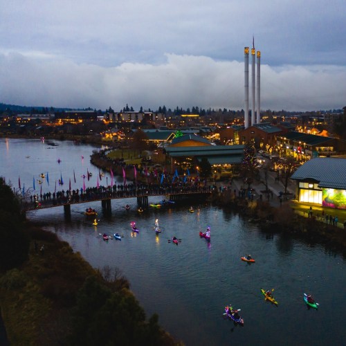 Aerial view of a river with illuminated kayaks and a crowd on a bridge, surrounded by buildings and cloudy skies.