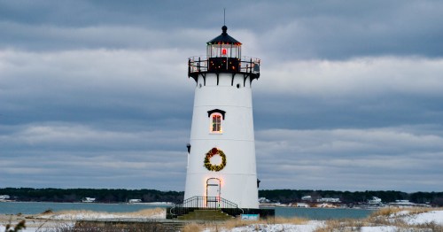 A white lighthouse adorned with a wreath, standing by the water under a cloudy sky.