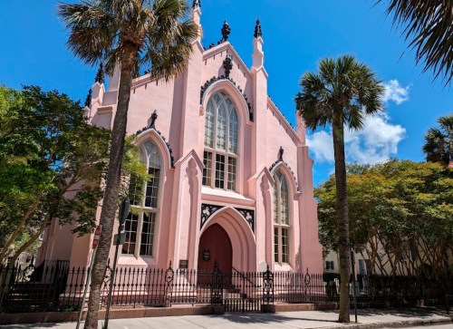 A pink Gothic-style church with tall windows and ornate details, surrounded by palm trees under a blue sky.
