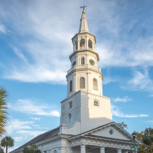 A tall white church tower with a clock, set against a blue sky with scattered clouds. Palm trees are in the foreground.