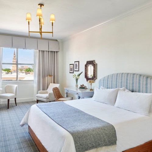 Bright hotel room featuring a large bed, elegant decor, a window with a view, and stylish furniture.