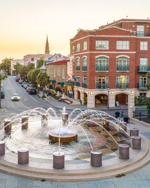A fountain with water jets in a plaza, surrounded by buildings and palm trees, under a sunset sky.