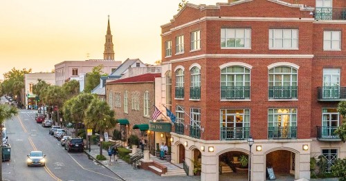 A charming street scene featuring a brick building, palm trees, and a church steeple in the background at sunset.