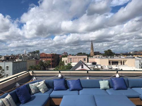 Rooftop view of a city skyline with clouds, featuring buildings and a church steeple in the distance.
