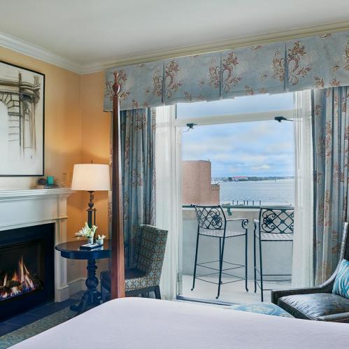Cozy hotel room with a fireplace, elegant decor, and a balcony overlooking a scenic waterfront view.