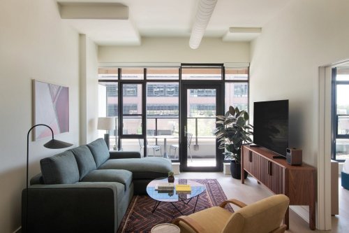 Modern living room with a green sofa, wooden TV stand, large windows, and a potted plant, featuring a cozy atmosphere.
