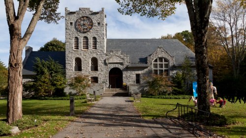 A stone building with a clock tower, surrounded by trees and a grassy area, under a clear blue sky.