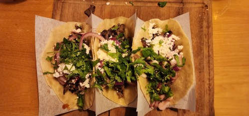 Three tacos filled with beef, onions, cilantro, and cheese on a wooden board.
