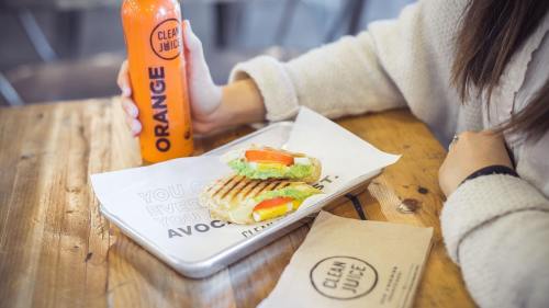 A person holds an orange juice bottle next to a plate with a grilled sandwich filled with vegetables on a wooden table.