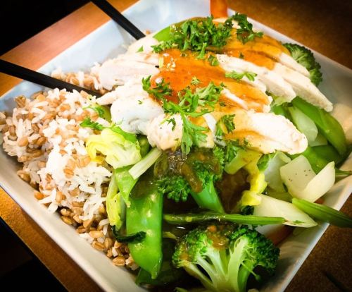 A bowl of rice and vegetables topped with sliced chicken and drizzled with sauce, garnished with herbs.