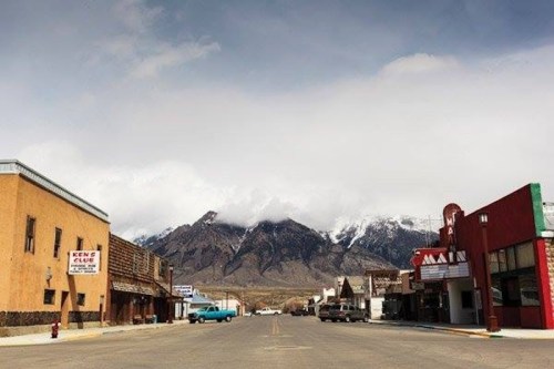 A quiet street in a small town with mountains in the background, featuring rustic buildings and cloudy skies.