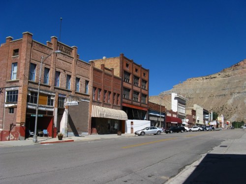 A quiet street lined with historic brick buildings and shops, set against a clear blue sky and rocky hillside.
