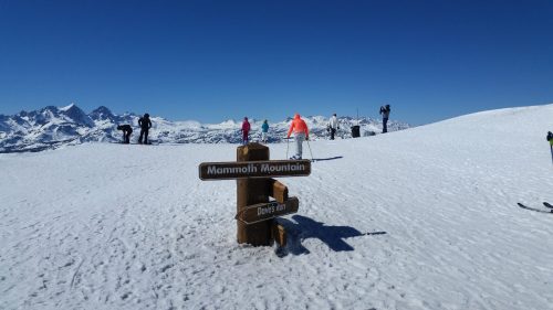 A snowy mountain landscape with skiers and a wooden sign pointing to Mammoth Mountain and Davis Run. Clear blue sky.
