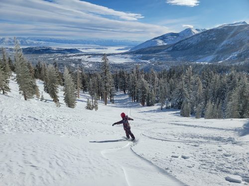 A child in a red snowsuit snowboards down a snowy slope, surrounded by trees and mountains under a clear blue sky.