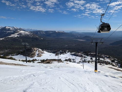 A snowy mountain landscape with ski lifts and a clear blue sky, surrounded by forested hills and distant peaks.