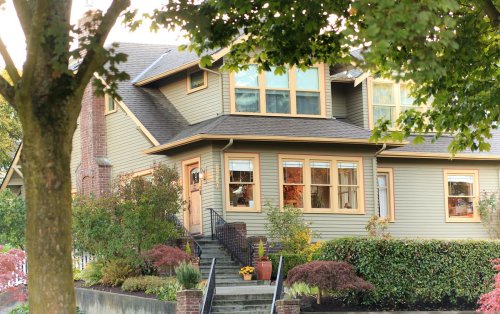A charming two-story house with green siding, large windows, and a landscaped front yard, framed by trees.