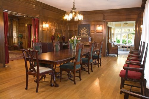 A spacious dining room with a large wooden table, elegant chairs, and rich wood paneling, leading to a sunlit area.