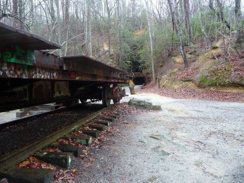 A rusted train car on tracks near a gravel path leading to a dark tunnel, surrounded by bare trees and rocky terrain.