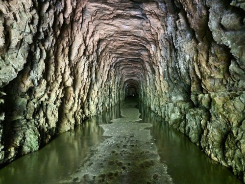A dimly lit underground tunnel with rocky walls and water on the floor, creating a mysterious atmosphere.