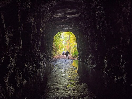 A dark tunnel with a view of two people walking towards light and greenery at the end, reflecting water on the ground.