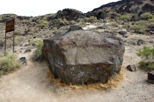 A large boulder with ancient petroglyphs, surrounded by dry grass and rocky terrain under a cloudy sky.