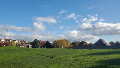 A grassy field with trees and buildings under a blue sky with clouds, featuring a distinctive conical structure in the background.