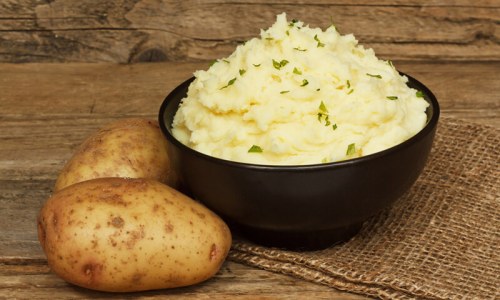 A bowl of creamy mashed potatoes garnished with herbs, next to two raw potatoes on a rustic wooden surface.
