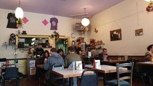 A cozy café interior with patrons dining, a counter with coffee equipment, and colorful decorations on the walls.