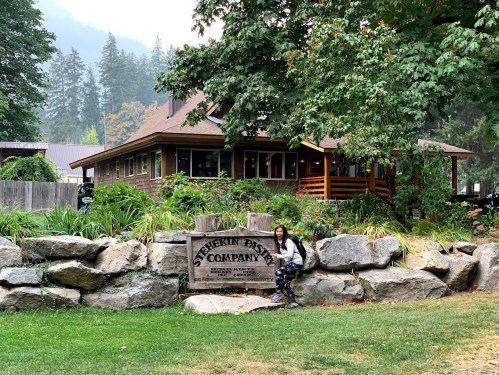 A woman sits on a rock near a sign for Stehekin Pastry Company, surrounded by greenery and a rustic building in the background.