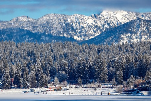 Snow-covered mountains rise above a forest of evergreen trees, with a frozen lake in the foreground.
