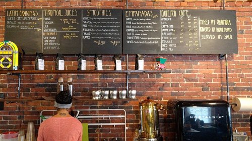 A person stands in front of a menu board featuring Latin food, tropical juices, smoothies, and empanadas in a café.