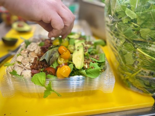 A hand adds toppings to a colorful salad with greens, tomatoes, and avocado, next to a large bag of fresh greens.
