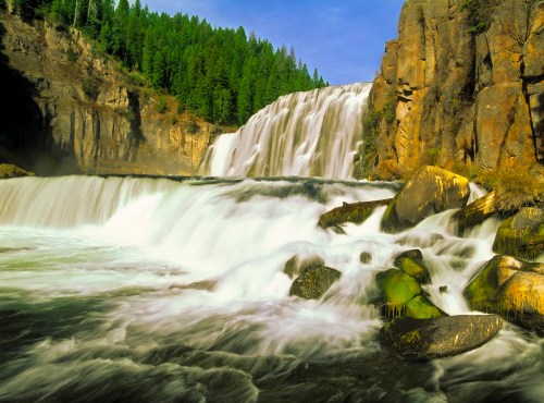 A cascading waterfall flows over rocky cliffs, surrounded by lush green trees under a clear blue sky.