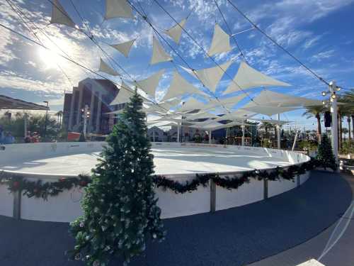 An outdoor ice skating rink decorated with holiday trees and white canopies under a bright blue sky.