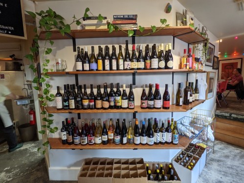 A wooden shelf displaying various bottles of wine, with plants and a cozy interior in the background.