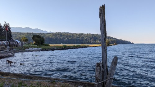 A serene lakeside view with calm water, distant mountains, and lush greenery under a clear blue sky.