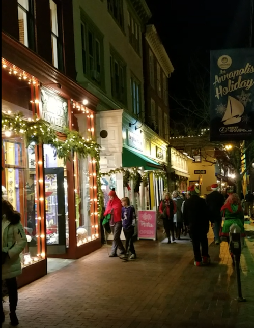 A festive street scene at night, with holiday lights, decorations, and people walking in Annapolis.
