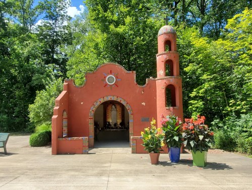 Colorful chapel structure surrounded by greenery, featuring a bell tower and vibrant flower arrangements in pots.