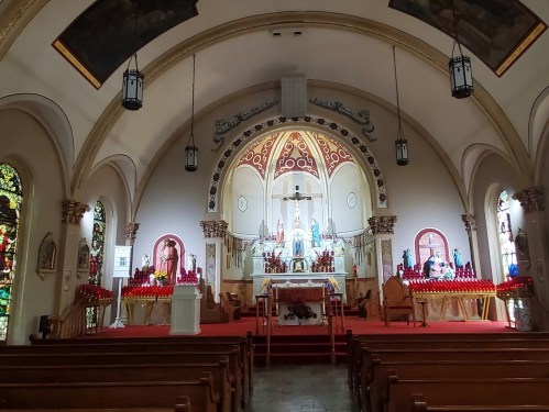 Interior of a church featuring an altar, stained glass windows, and wooden pews, illuminated by soft lighting.