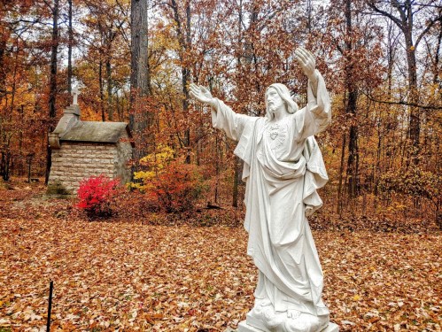 A white statue of Jesus with outstretched arms stands in a forest of autumn leaves, near a small stone chapel.