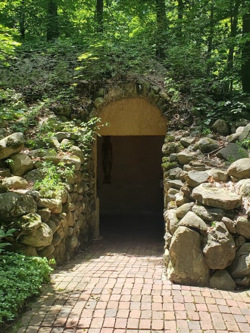 A stone archway leads into a wooded area, with a path of bricks and greenery surrounding the entrance.